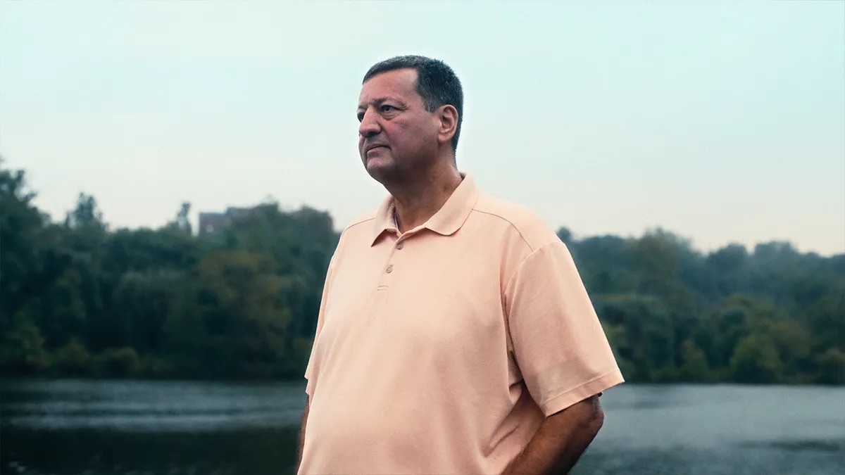 Bruce Plaxen attorney profile thumbnail. A man in a peach polo shirt stands beside a lake with trees reflected in the water.