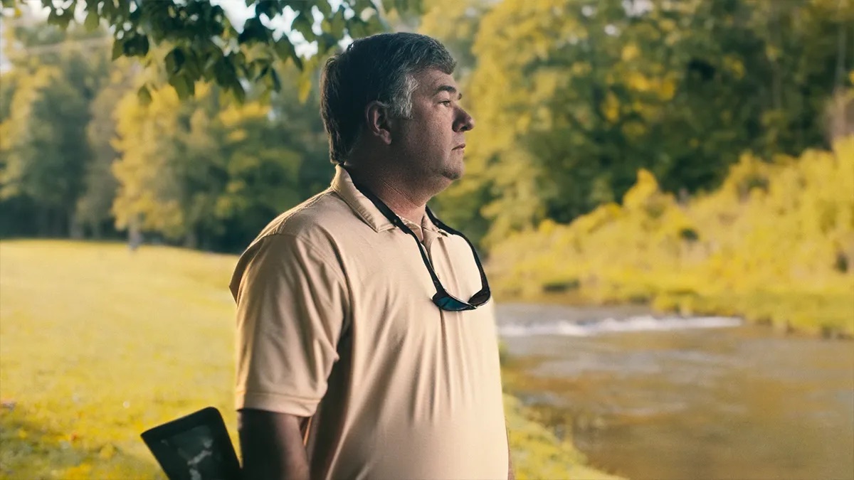 Clint Scott attorney profile thumbnail. A man with greying hair in a tan polo shirt, sunglasses hanging from the collar, standing in profile beside a creek with bright green trees.