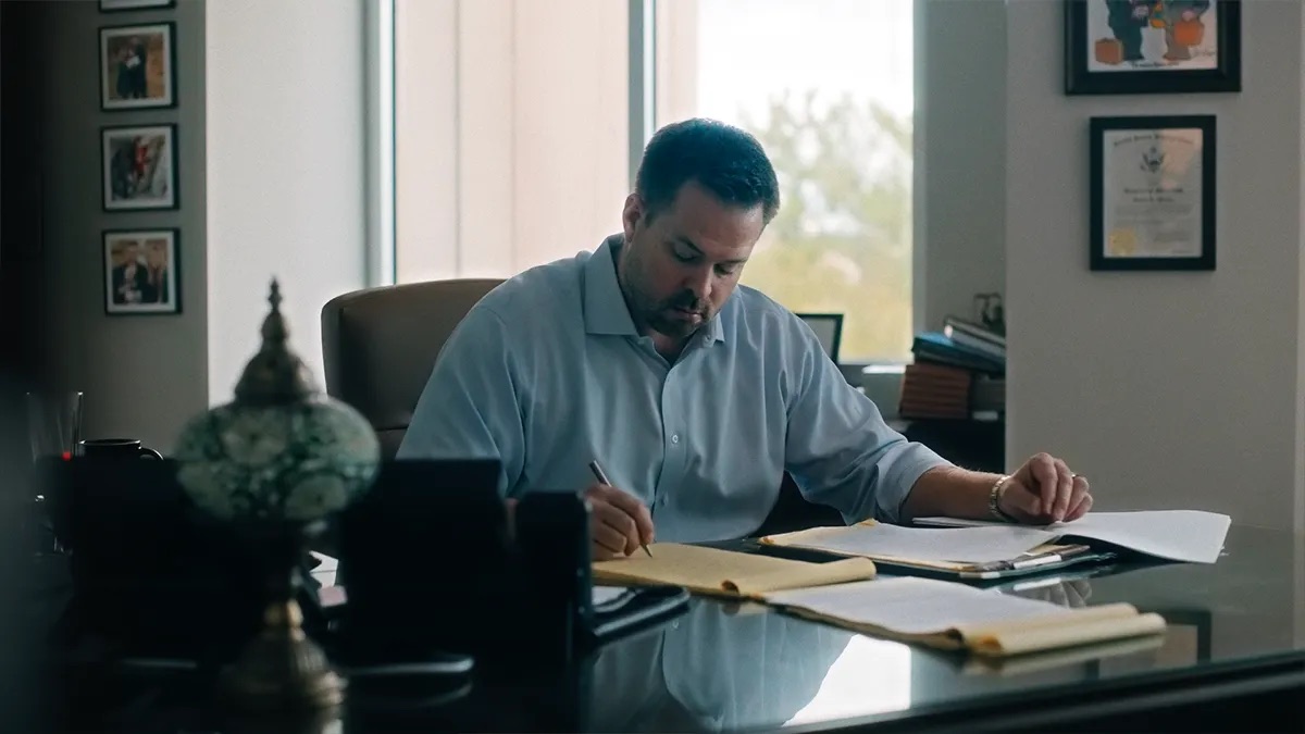 David Muncy attorney profile thumbnail. At a dark desk writing with a pen, yellow legal folders and white papers in front of him, framed photographs and a certificate on the walls.