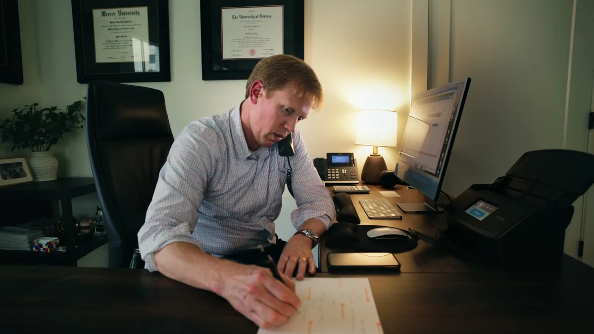 Kevin Patrick attorney profile thumbnail. Phone to his ear, writing on a notepad, a man in a blue gingham dress shirt at his desk with framed diplomas and a lamp behind him.
