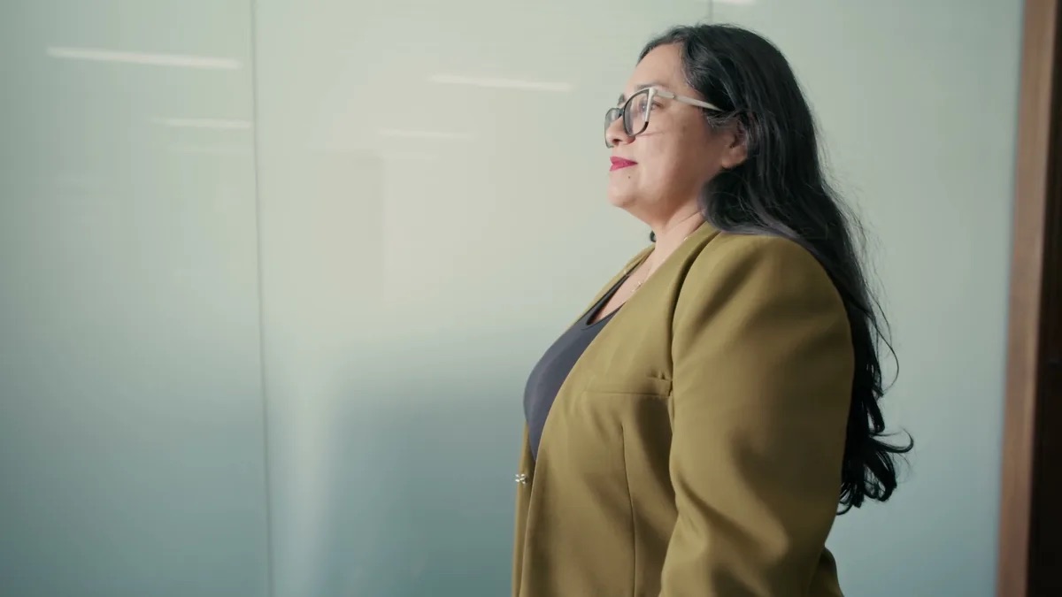 Vilma Duarte Spanish-language attorney profile thumbnail. Indoors beside a frosted glass partition, a woman in a mustard blazer and large glasses.