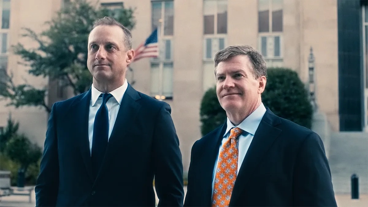 Cunningham & Mears brand film thumbnail. Two men in dark suits stand outdoors in front of a tan stone building with an American flag on a pole and some shrubs.