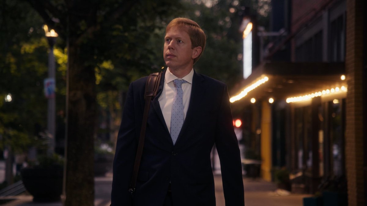 A man in a dark suit with a shoulder bag walking along a tree lined sidewalk at dusk, string lights glowing over storefronts beside him