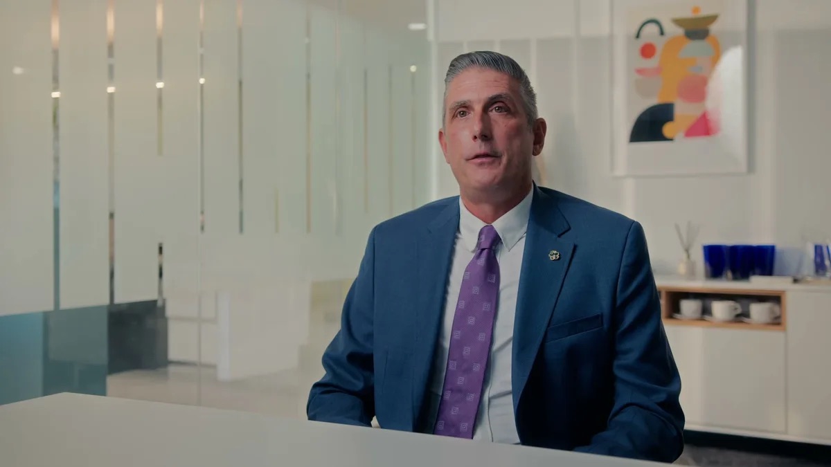 Padula Law brand film thumbnail. A man in a navy suit and purple tie sits at a desk with a white credenza behind him.