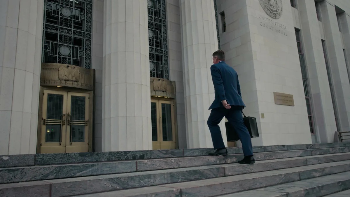 KMD Law FAQ video thumbnail on going to trial. Seen from behind climbing marble steps, an attorney in a blue suit with a briefcase, and the stone facade carved UNITED STATES COURT HOUSE.