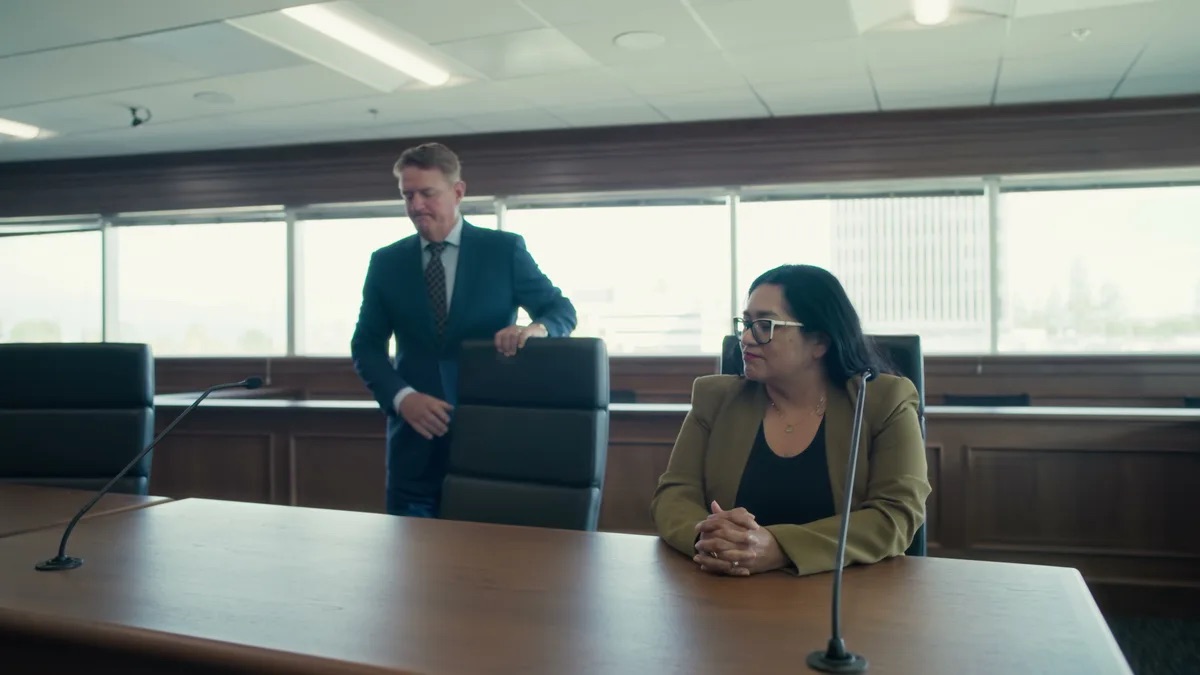 KMD Law FAQ video thumbnail on seeing the perpetrator. A man standing behind a chair with his hand on its back in a dark suit, a woman seated at the long table with her hands folded.