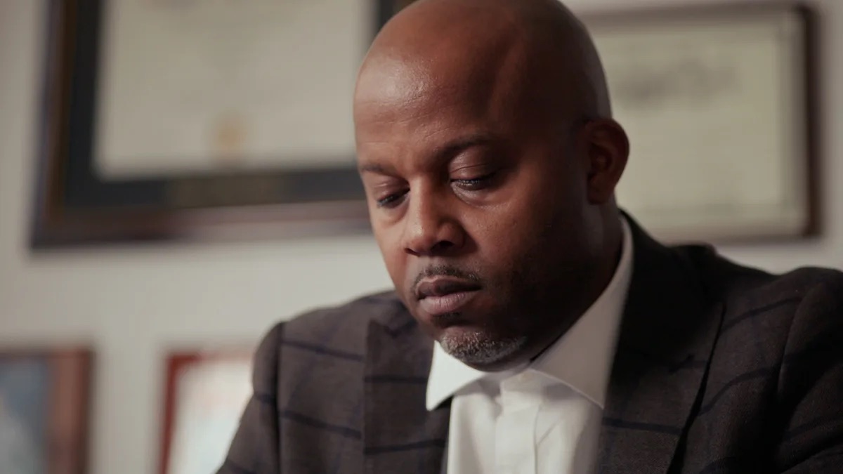 Lessem, Newstat & Tooson practice area video thumbnail on civil litigation. A man in a gray suit with a windowpane check looks down, framed certificates on a wall behind him, out of focus.