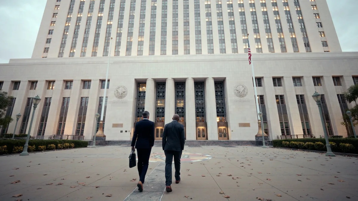 Lessem, Newstat & Tooson practice area video thumbnail on criminal defense. Seen from behind, two men in dark suits walking toward a pale stone building with fluted columns and an American flag.