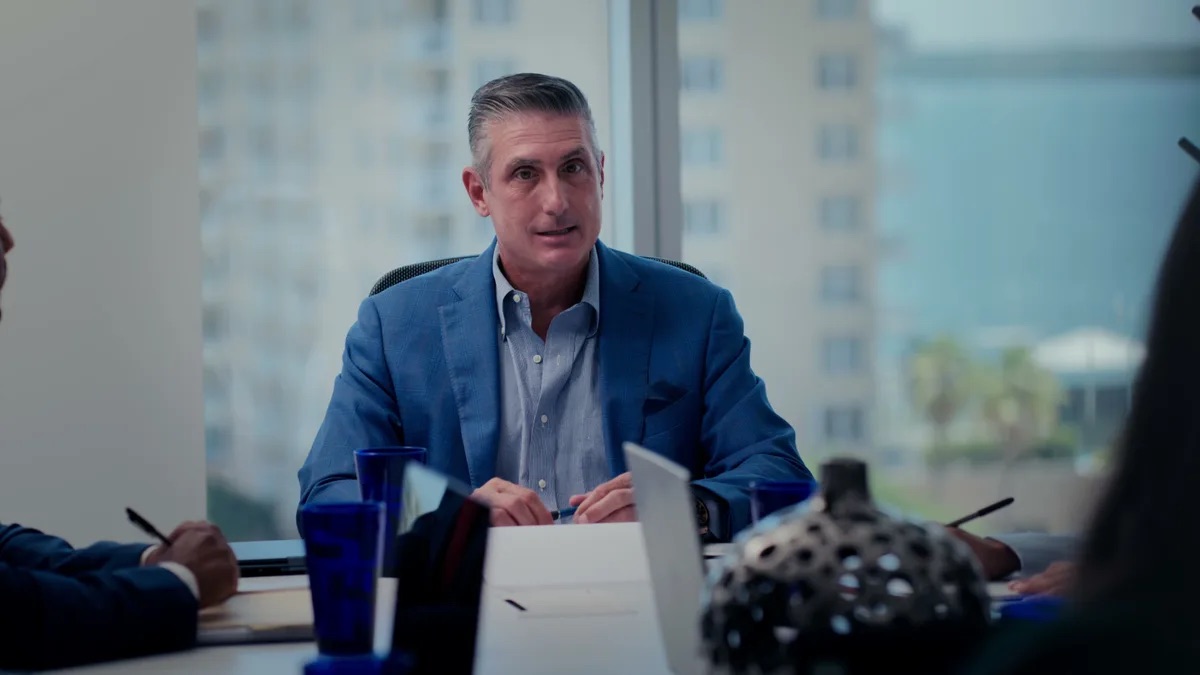 Padula Law practice area video thumbnail on asset forfeiture. An attorney in a blue blazer sits across a conference table from two people, cobalt blue drinking glasses and papers on the table.
