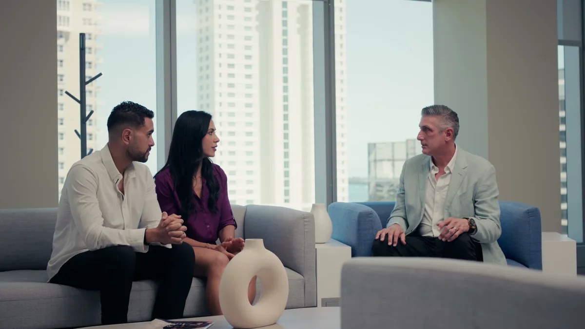 Padula Law practice area video thumbnail on criminal defense. A man and a woman on a gray sofa and a man in a light gray blazer facing them on a blue sofa, tall windows with towers outside.