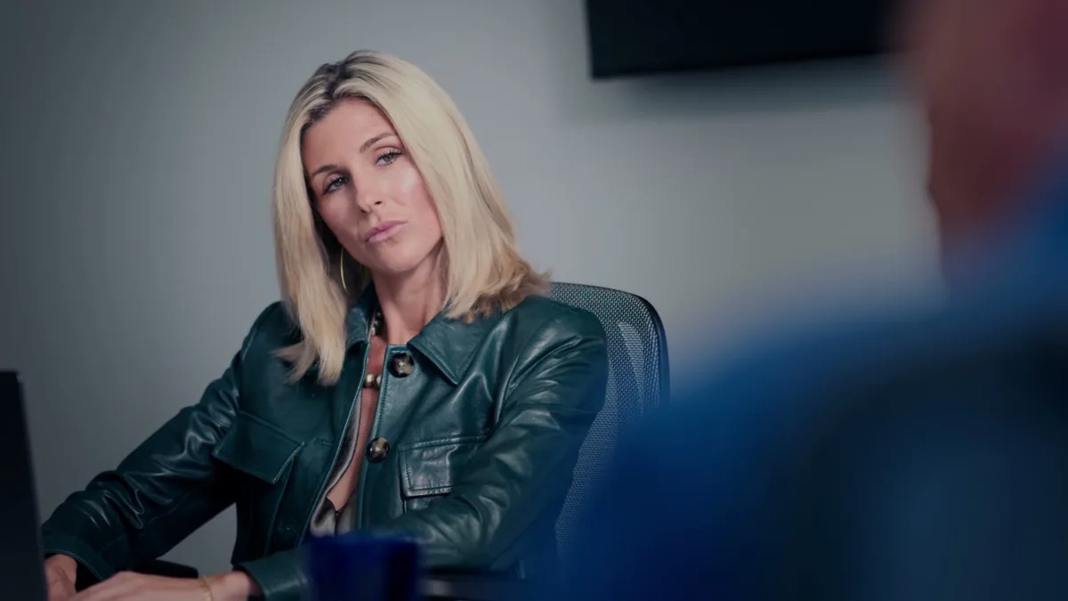 Padula Law practice area video thumbnail on fraud advocacy. A blonde woman in a dark green leather jacket at a desk, looking toward someone out of frame, a dark screen on the wall behind her.