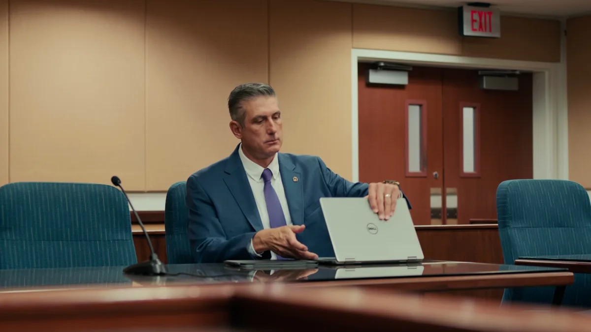 Padula Law practice area video thumbnail on healthcare fraud. An attorney in a blue suit sits at a long dark table with a gooseneck microphone, teal chairs, an EXIT sign over dark red double doors.