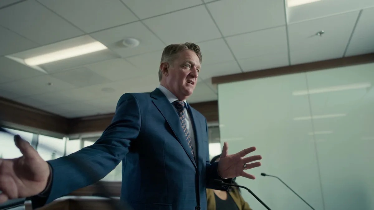 KMD Law practice area video thumbnail on personal injury. An attorney in a blue suit gestures with both arms spread wide, shot from below, a frosted glass partition behind him, microphones.