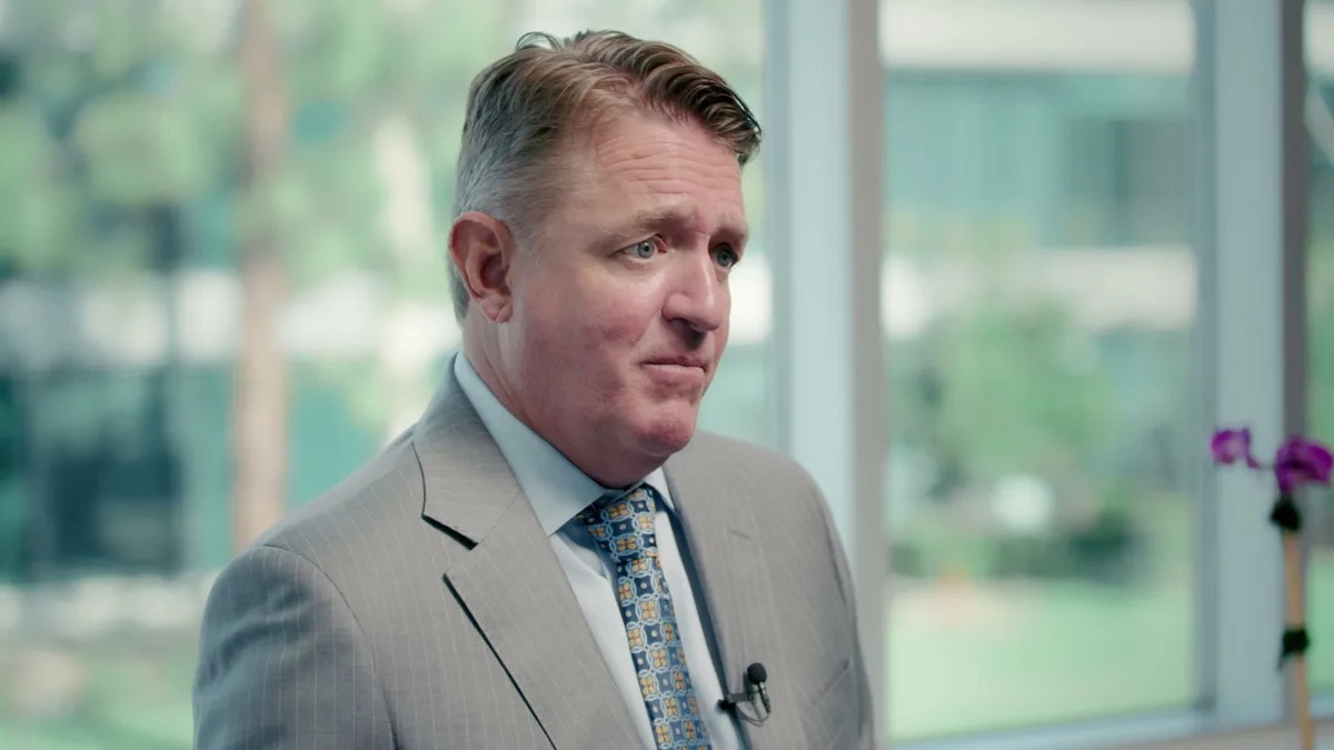 KMD Law practice area video thumbnail on sexual assault. An attorney in a light gray suit stands beside a wall of windows with green trees outside, a small lavalier mic clipped to his tie.