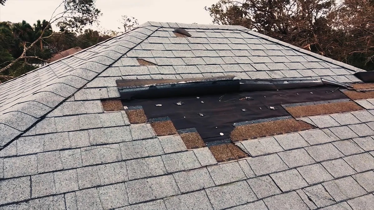 Practice area video thumbnail on wind damage. A damaged roof with missing shingles and exposed black underlayment, trees in the soft background.