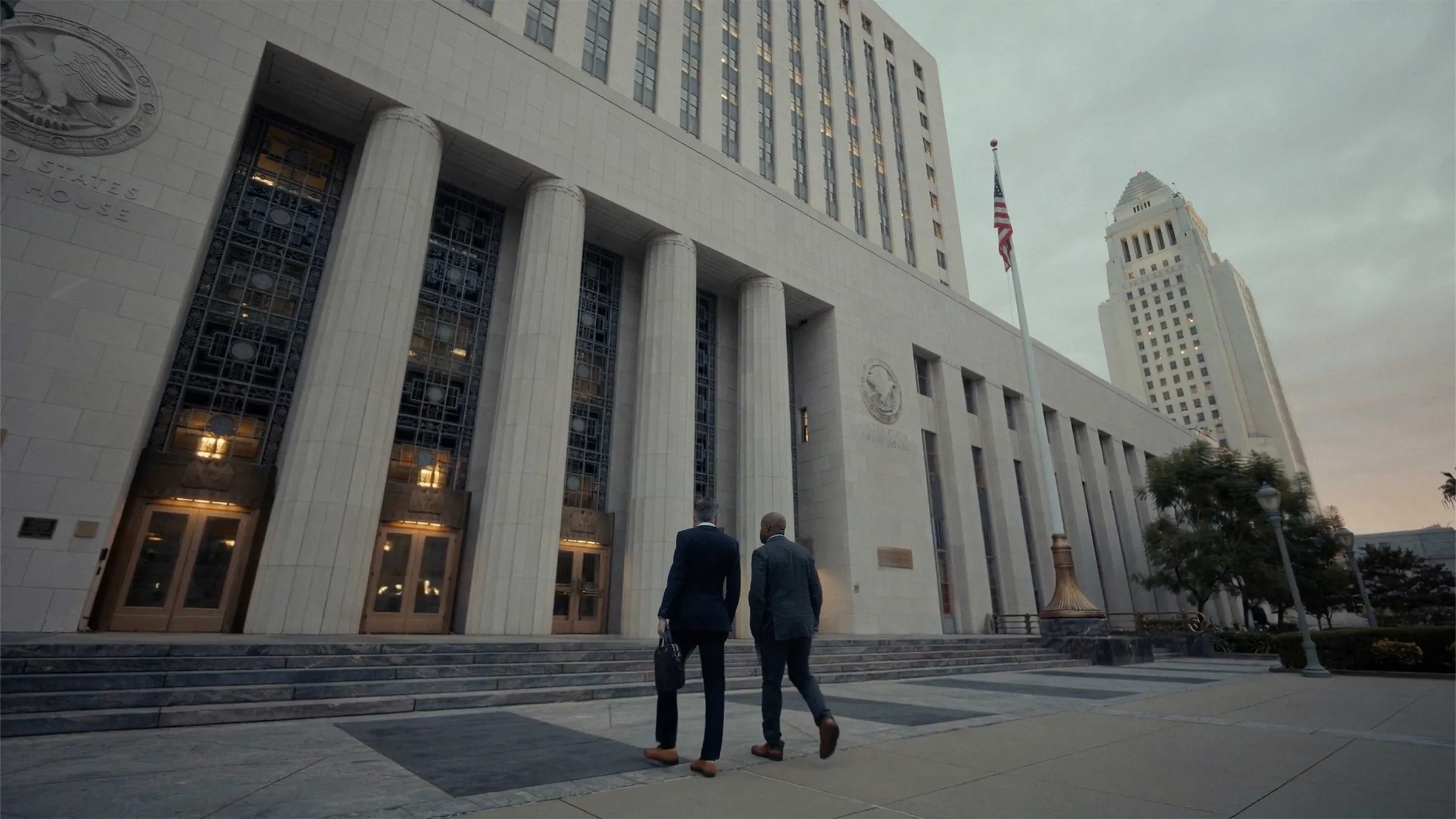 Two men in suits walk toward a pale stone building with columns and a United States Court House inscription, an American flag between the building and a tall stepped tower on the right